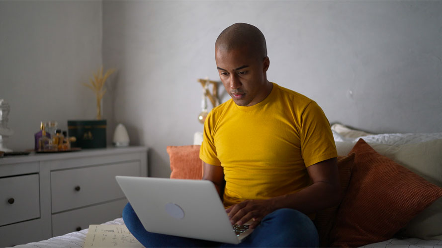 hombre joven de color con camisa amarilla usa una computadora sentado en la cama