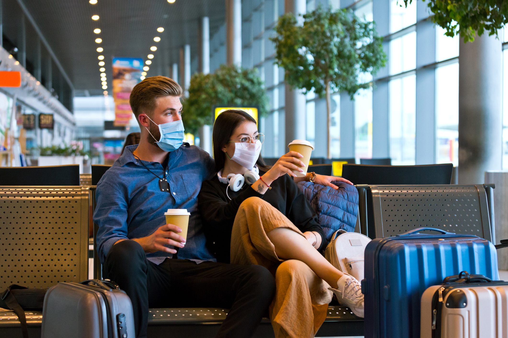 Mujer joven y hombre viajando en avión durante la pandemia por COVID-19 con mascarillas faciales N95, sentados en un banco con un café en el área de espera del aeropuerto.