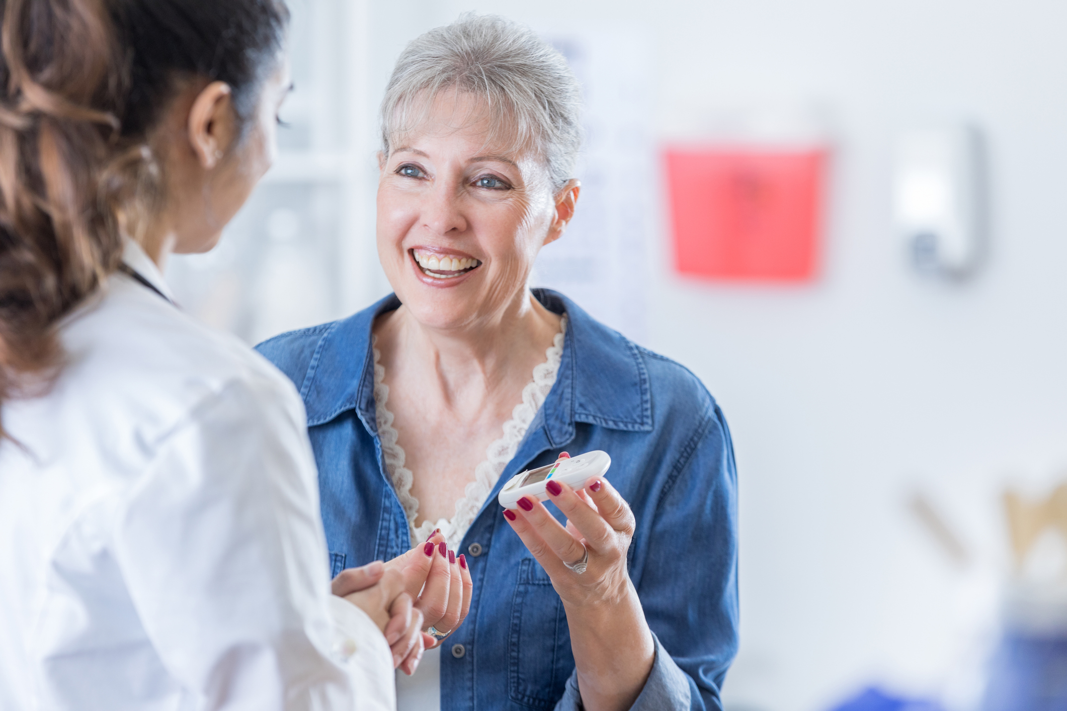 Una señora mayor sonriente sostiene un glucómetro para controlarse su nivel de azúcar en la sangre. Está hablando con su médico.