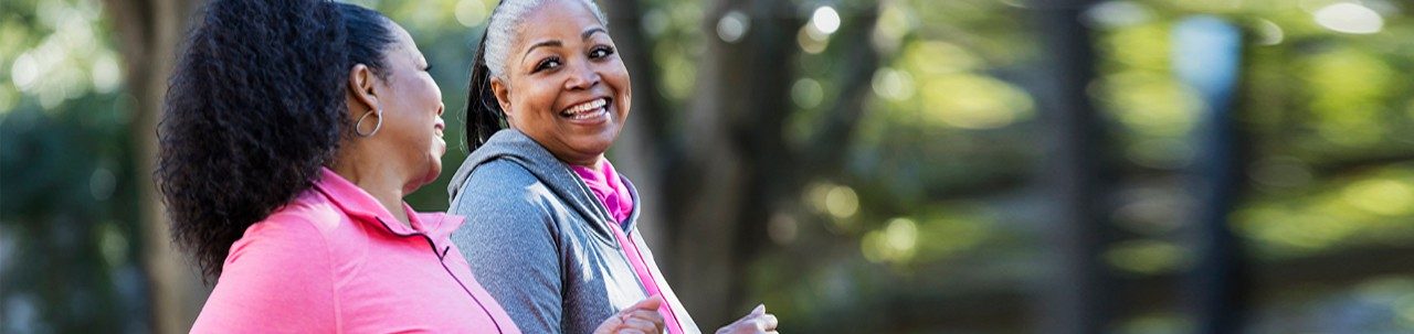 Dos mujeres afroamericanas haciendo ejercicio juntas en la ciudad, trotando o caminando, riendo y conversando. Los edificios y árboles están fuera de foco en el fondo. La de color rosa tiene 60 años y su amiga tiene 50.