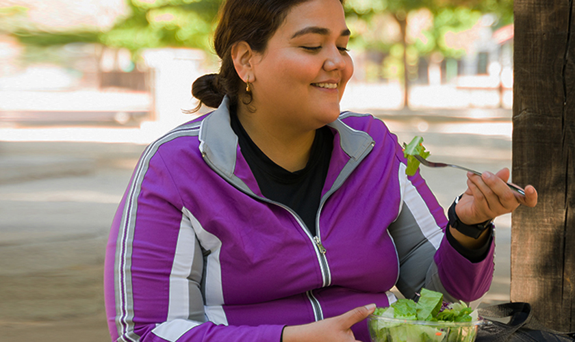 Mujer feliz con sobrepeso sonriendo mientras come una ensalada verde saludable antes de comenzar sus ejercicios aeróbicos al aire libre